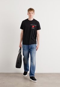 Zwarte katoenen T-shirt met grafische logo, gecombineerd met blauwe denim jeans. Model houdt een zwarte tote bag vast en draagt zwarte sneakers.