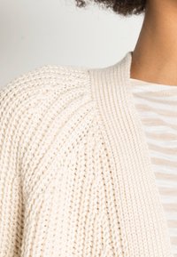 Knit cardigan vaaleassa värissä, jossa on muodostettuja ribbitekstuureja ja rento istuvuus, yhdistettynä raidalliseen vaaleaan paitaan alla.