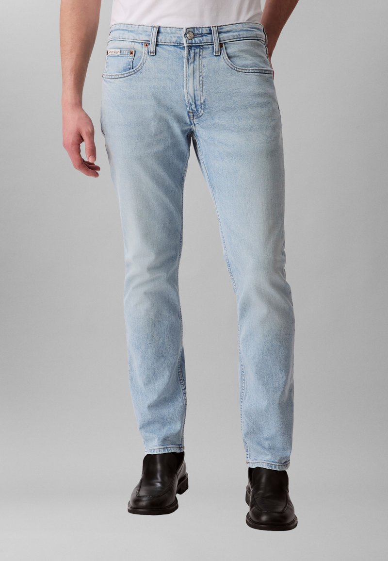 Gaiši zili džinsu džinsi ar taisnu piegriezumu, piecu kabatu dizainu un smalku izbalējumu, kombinēti ar melniem ādas apaviem.