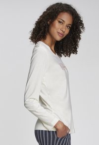 Langärmliges cremefarbenes Oberteil mit lockerer Passform, Rundhalsausschnitt und korallfarbenem Grafikdesign auf der Vorderseite. Weiche Stofftextur und lässiger Stil.