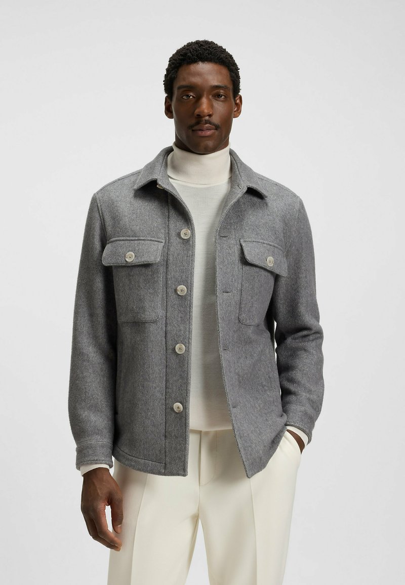 Gray wool blend jacket with a herringbone pattern, two chest pockets, button closure, and a structured collar. Worn over a light turtleneck.