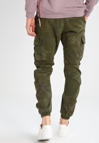 Zielone spodnie bojówki w kamuflażu z elastycznymi mankietami, wieloma kieszeniami i gładką teksturą materiału, zestawione z białymi trampkami.