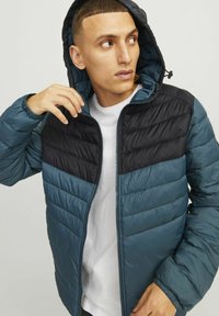 Chaqueta acolchada en azul verdoso oscuro y negro, con cremallera frontal, capucha y costuras en forma de chevron en el pecho. Textura de tela suave y brillante.