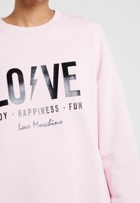 Person som bär en ljusrosa sweatshirt med ordet "LOVE" i svart, där en blixt ersätter "O", och mindre text nedanför.
