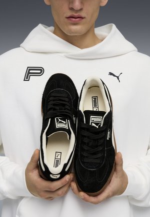 Sorte sneakers med overdel af suede og tekstil, med hvide detaljer og en struktureret gummisål. Holdt af en person i en hvid hættetrøje.