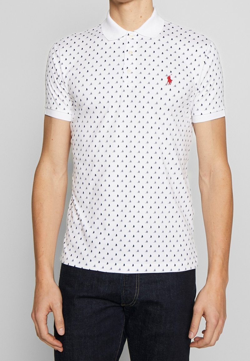 Homme portant un polo blanc avec de petits motifs de voiliers bleu marine et un logo rouge sur la poitrine, associé à un jean bleu foncé.