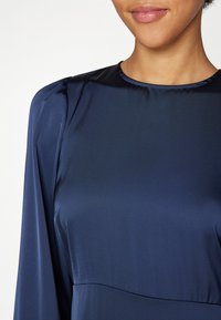 Navy sininen satiinimekko, jossa on pitkät hihat, pyöreä kaula-aukko ja hienovaraiset poimutustiedot olkapäissä, sekä sileä pinta.