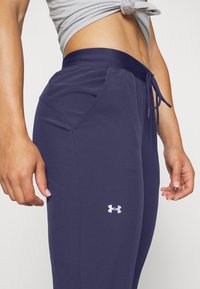 Närbild på en person som bär marinblå Under Armour leggings med sidofickor och dragsnodd i midjan, som visar en del av deras mage och händer.