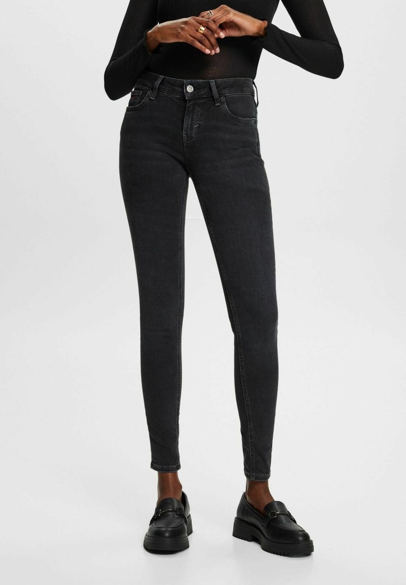 Personne portant un jean noir ajusté, un haut noir à manches longues et des mocassins noirs, debout avec les mains jointes à la taille sur fond blanc.