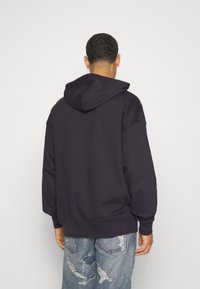 Hombre de espaldas vistiendo una sudadera negra con capucha y jeans claros rasgados frente a un fondo blanco liso.