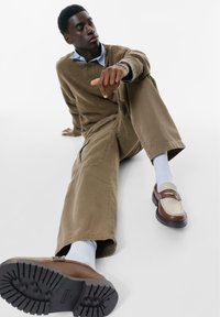 Pulover tricotat maro, cămașă cu guler albastru deschis, pantaloni verde măsliniu și mocasini bicolori. Sosetele sunt albe. Tălpile pantofilor au o textură cu aderență.