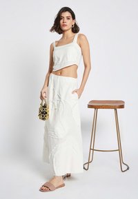 Weißes Leinen-Crop-Top mit quadratischem Ausschnitt, kombiniert mit einem fließenden Maxirock mit strukturierten Mustern und einem fransigen Saum. Kombiniert mit einer kleinen schwarzen und goldenen Tasche sowie flachen Sandalen.