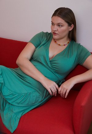 Vestido verde em um tecido suave e elástico, com uma bainha larga e faixa na cintura. Usado com um colar de contas coloridas, sentado em um sofá vermelho.