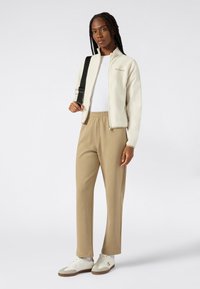 Giacca bianca con zip e logo, abbinata a pantaloni della tuta beige e sneakers color crema. Texture liscia, design aderente e cintura elastica.