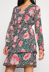 Kukkamekko mustalla taustalla, jossa on vaaleanpunaisia ruusuja ja vihreitä lehtiä. Pitkät hihat, rypytetty vyötärö ja kerroksellinen hameen muoto.