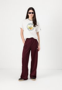 Tricou alb cu logo circular Adidas, asociat cu pantaloni de trening maro, largi, care au dungi laterale și logo. Pantofi negri slip-on.