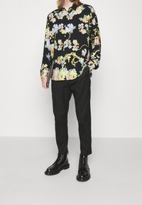 Camicia nera a maniche lunghe con motivi floreali colorati. Abbinata a pantaloni neri e stivaletti neri con lacci. Tiene un tablet.