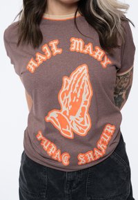 Brown graphic t-shirt featuring orange "HAIL MARY" text and a praying hands illustration. Round neckline with beige accents. Short sleeves.