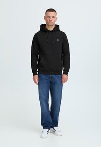 Svart hettegenser med snøring, frontlomme og brodert logo; kombinert med blå jeans og hvite treningssko.