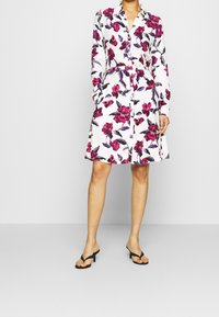 Donna che indossa un vestito camicia bianco floreale con fiori rosa e viola, maniche lunghe, cintura e sandali neri con tacco, in piedi su uno sfondo semplice.