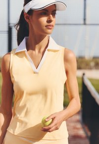 Femme en tenue de tennis jaune sans manches et visière blanche tenant une balle de tennis sur un court de tennis extérieur.