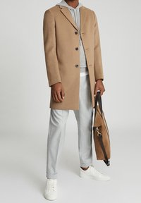 Man in een beige jas over een grijze hoodie en lichtgrijze pants, die een tan suède tas met zwarte straps vasthoudt, staand op een effen lichte achtergrond.