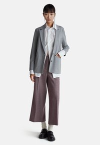Šedý přizpůsobený blazer přes bílou košili, spárovaný s vysokým pasem, širokými kalhotami s kostkovaným vzorem a černými mokasínami, vytváří moderní vzhled.