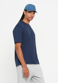 Mlada osoba s dugim pletenicama nosi plavu kapu, tamnoplavu majicu i svijetlosive trenirke, stoji na jednobojnoj bijeloj pozadini.