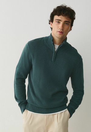 Ung mand iført en mørkegrøn striktrøje med kvart lynlås og beige bukser, stående med hænderne i lommerne mod en ensartet lys baggrund.