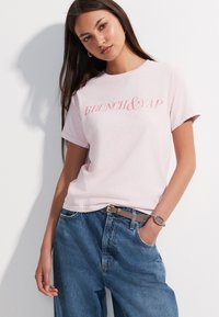 Svijetloružičasta pamučna majica s natpisom "BRUNCH & YAP", ležerni kroj, kratki rukavi, kombinirana s opuštenim plavim trapericama i smeđim remenom.