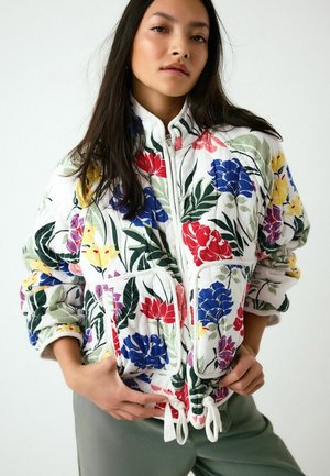 Frau trägt eine weiße Steppjacke mit roten, blauen, gelben und lila Blumenmustern, steht mit den Händen nahe den Taschen vor einem einfarbigen Hintergrund.