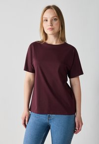 Burgunderfarget bomulls t-skjorte med korte ermer, rund hals, avslappet passform og sidesplitter, sammen med blå jeans mot en nøytral bakgrunn.