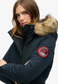 Dark navy puffer jacket with a faux fur hood, zippered front, and a red circular patch featuring mountains on the left sleeve.