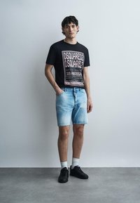 Νέος άνδρας φοράει μαύρο graphic t-shirt DSQUARED2, ανοιχτό μπλε τζιν σορτς, λευκές κάλτσες και μαύρα sneakers, στέκεται πάνω σε έναν απλό τοίχο.