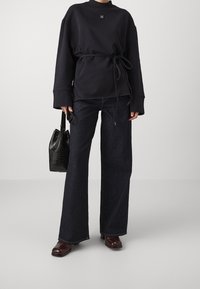 Persona che indossa una maglietta nera a maniche lunghe con cintura in vita, jeans ampi scuri, scarpe marroni e tiene in mano una borsa nera testurizzata.