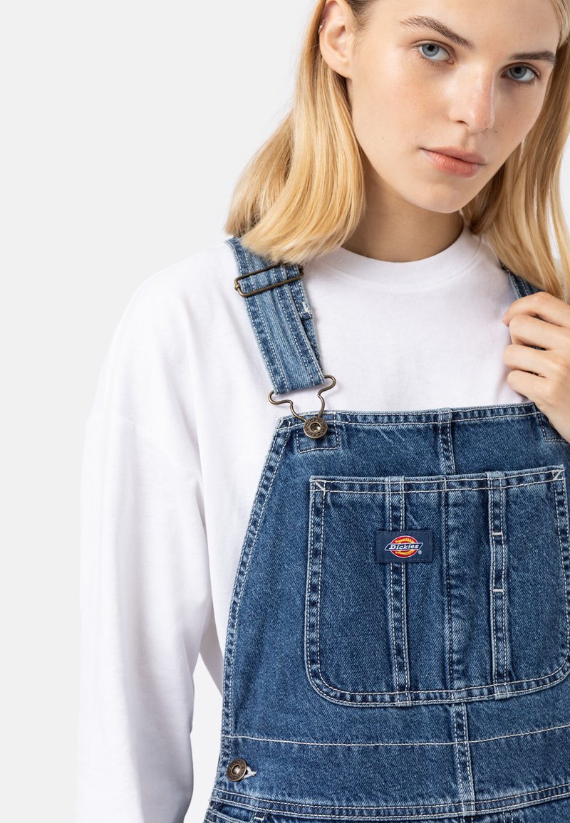 Salopette en denim bleu moyen avec des bretelles réglables, une poche avant et un patch logo. Superposée sur une chemise à manches longues blanche. Texture lisse.