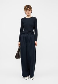 Γυναίκα με μαύρο μακρυμάνικο τοπ και μπλε παντελόνι με φαρδιές πιέτες, κρατώντας σκουρό καφέ τσάντα, στέκεται μπροστά σε λευκό φόντο.