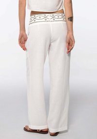 Pantalones de lino blancos de pierna ancha con una cinturilla decorativa y estampada que presenta acentos metálicos. Textura suave, corte holgado, longitud hasta el tobillo.