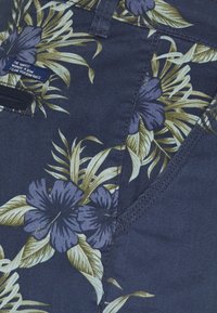 Miehen tummansininen kangas, jossa on kukkakuosi, jossa on violetteja ja vihreitä lehtiä. Lähikuvassa näkyy ompelutiedot ja pieni tagi.