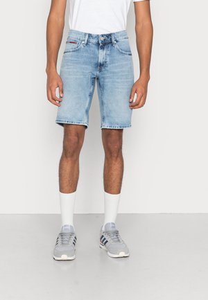 Mand iført lyseblå denimshorts, hvide sokker, grå løbesko og en hvid t-shirt, der står mod en ensfarvet baggrund.