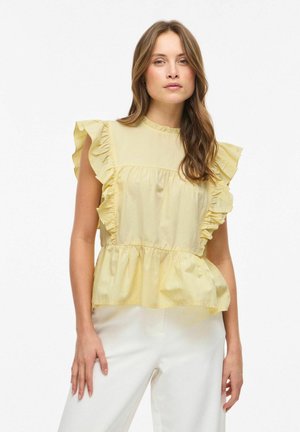 Kvinde med langt brunt hår iført en lysegul rynket ærmeløs bluse og hvide bukser, stående foran en ensfarvet hvid baggrund.