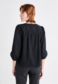 Black blouse with textured dot pattern, puffed sleeves, and lace trim at cuffs. Slightly flared shape, button closure at back.