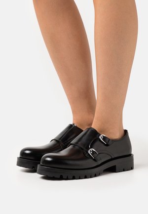 Zapatos de cuero negro con puntera redondeada, que presentan dos hebillas de plata. Suela de goma resistente con un patrón de textura.