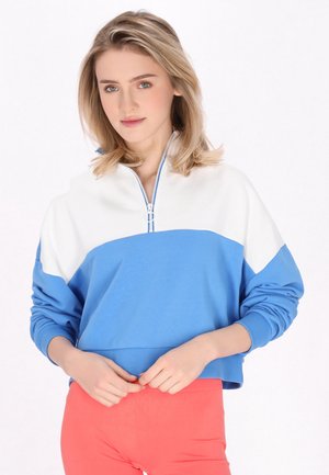 Ung kvinde med blondt hår iført en blå og hvid lynlås-sweatshirt og koralfarvede bukser, stående med hænderne nær taljen mod en hvid baggrund.
