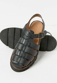 Black leather sandals with woven design, open back, adjustable buckle, and rubber sole with textured grip. Notable stitching detail around the sole.