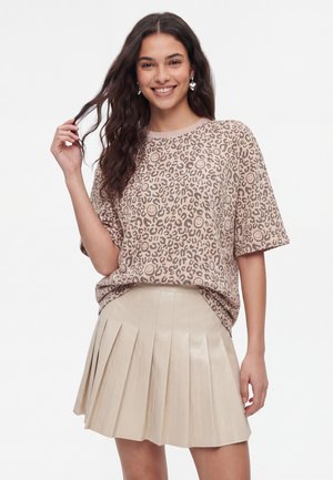 Kvinde med langt mørkt hår iført en beige leopardprint t-shirt og et lys beige plisseret nederdel, smilende og rører ved sit hår.