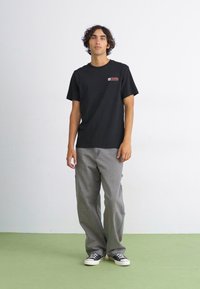 Black short-sleeve t-shirt with a small red and white logo on the chest, paired with gray wide-leg pants and black sneakers.