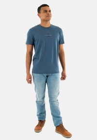 Camiseta de algodón azul con mangas cortas y un pequeño detalle gráfico en el pecho, combinada con jeans azul claro y zapatos marrones.