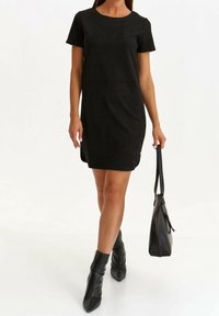 Robe noire à manches courtes avec un col rond, texture lisse, longueur au-dessus du genou, assortie d'un sac à main noir et de bottes à chevilles.