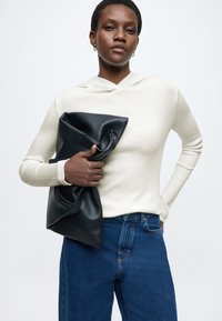 Világos színű bordázott pulóver, kék farmernadrág és egy fekete bőrborítású, puha tapintású clutch táska, laza fogással tartva.